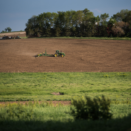 A tractor pulling a planter, planting in a bare field of brown soil. With a grassy area in the foreground and a tree line on the other side of the field.