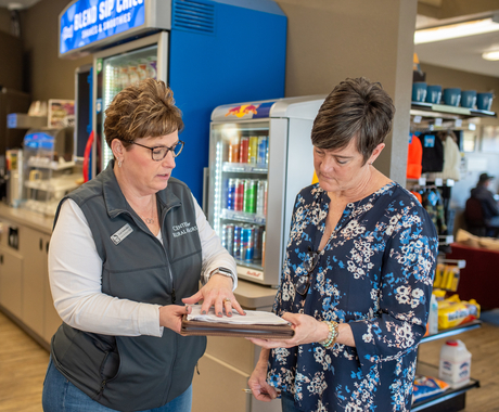 Two white women with short brown hair stand inside a convenience store with a large blue machine and a beverage cooler in the background while looking at papers