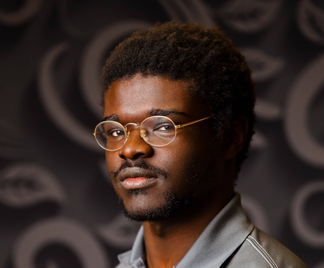 Peter David, a young African American wearing round gold framed glasses poses por a picture in front of a dark background wearing a grey polo shirt with a red and black logo on left side of shirt 