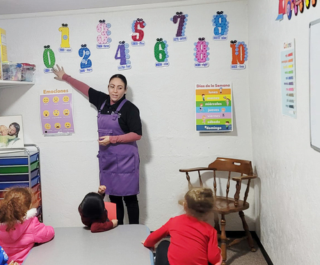 Maestra al frente del salón señalando algo en la pared a los niños sentados junto a una mesa