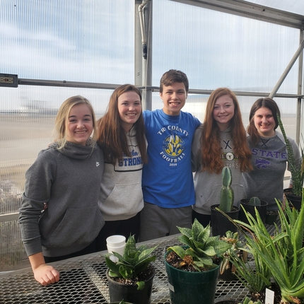 Five teenagers, two girls on the left, boy in the middle, two girls on the left. Plants on a table in front of them.