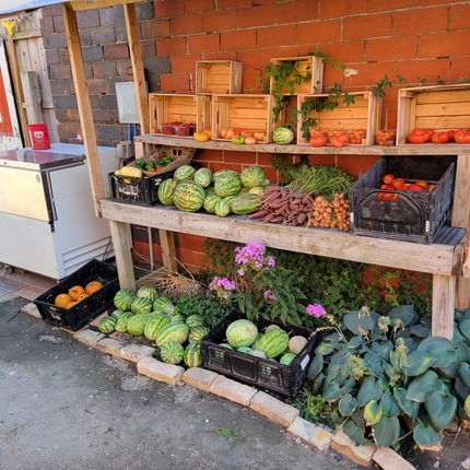 A vegetable and fruit stand filled with a variety of fresh produce, with six small wooden crates above also with produce against a red brick wall
