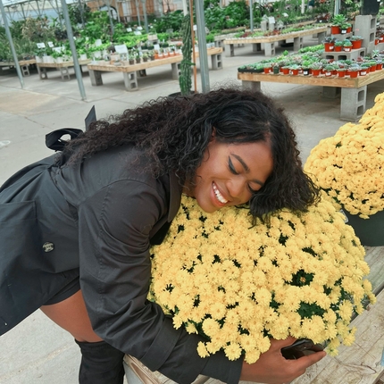 African American woman wearing a long sleeve jacket and high black boots hugs a pot of yellow flowers in a greenhouse