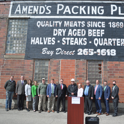 A group of people in front of a building and behind a podium, underneath a sign that says "Amend's Packing Plant"