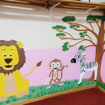 Mural de dibujos con un león, mono y una cebra y un árbol en una pared color rosa y blanco