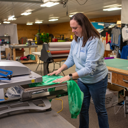 White woman with shoulder length brown hair wearing a light blue hoodie sweatshirt and blue jeans inserts a green shirt into a machine
