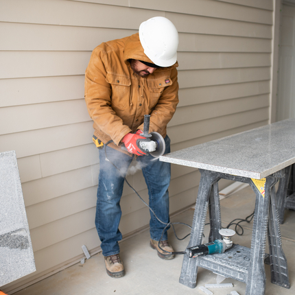 Man in think brown coat, blue jeans, work boots and a white hard hat cuts granite with a tool.
