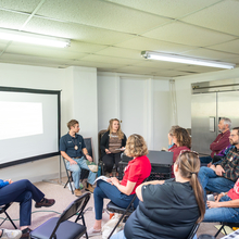 Diverses persones seuen en cadires en una sala amb poca llum escoltant una taula rodona de quatre persones.