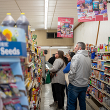 Three people stand in the aisle of a small grocery store looking at items on the shelves.