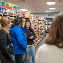 Several people stand in a grocery store aisle as one woman speaks.
