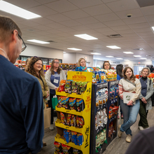 Several people stand tightly together in a small grocery store aisle listening to someone speak.