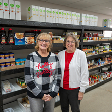 Two white females stand next to shelves of food in a food pantry.
