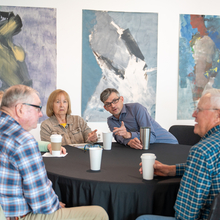 Una mesa con cinco personas conversando. Están frente a grandes obras de arte en la pared.