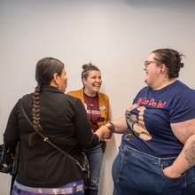 Three women laugh together, one woman with a long braid is looking away from the camera while the other two women look at each other with smiles