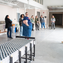 A group tours a distribution area, looking at a table on wheels, led by a woman on the opposite side of the room holding a paper