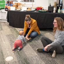 Two women sit crosslegged on the floor in front of a table with a black tablecloth, a baby crawls toward one of the women