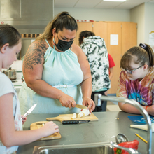 Woman cuts food as two students look on