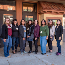 Group of eight women stand in front of a business on a sidewalk for a picture