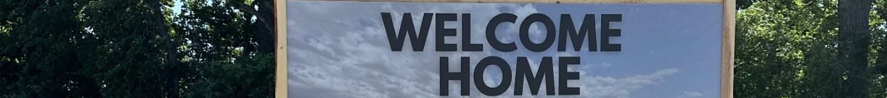A temporary sign that says "Welcome Home" it's on a dirt patch with sparse green grass with leafy trees in the background