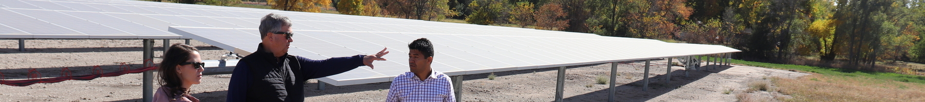 Three individuals - one woman and two men - stand in front of a solar array with brown grass in the foreground and trees in the background. The center man is pointing to the array.