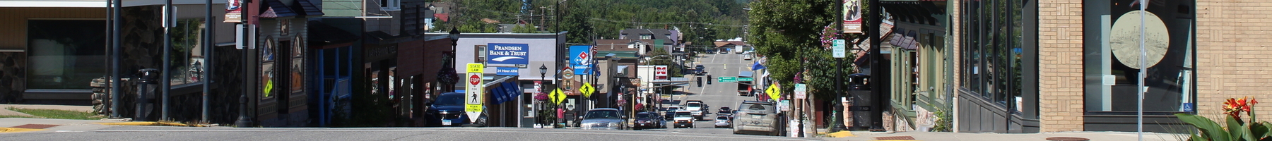 Small town main street with blue skies and puffy white clouds. A watertower is in the background, and a tree-filled landscape is behind that