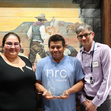 Photo of three people standing in front of a cowboy mural. The one in the center is holding an award.