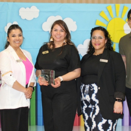 An image of five people standing in front of a blue-sky cutout. The woman in the middle is holding an award.