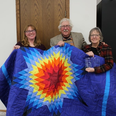 An image of three people standing holding a brightly colored quilt. The woman on the far right is holding an award.