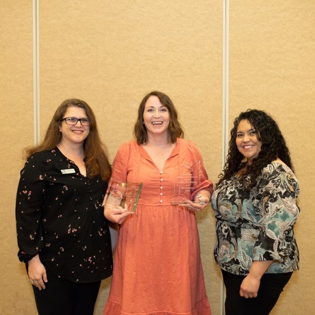 Three people standing in front of an orange background. The woman wearing orange in the center is holding an award