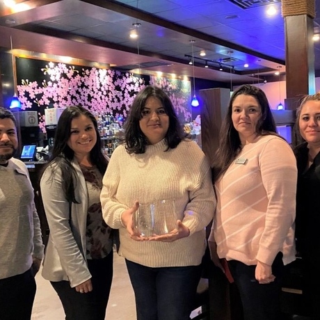 An image of five people standing in a restaurant. The woman in the middle is holding an award.