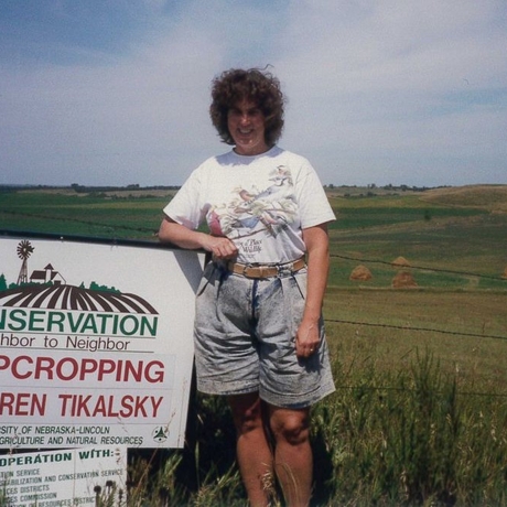 A woman standing by a conservation sign that reads "STRIPCROPPING JIM/KAREN TIKALSKY and in front of a field