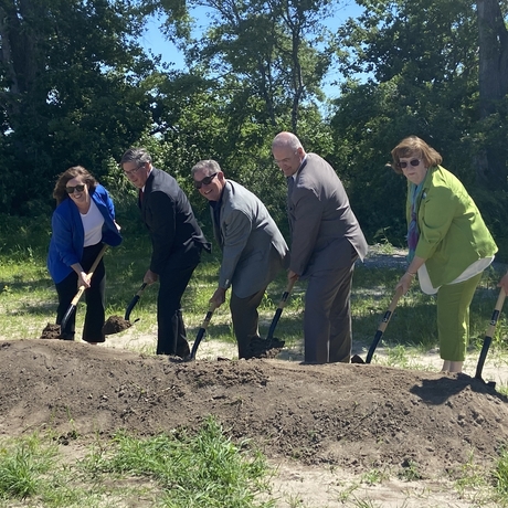 Six people in business clothes holding shovels, scooping up a mound of dirt in a ground breaking.