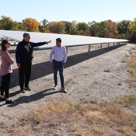 Tres persones, una dona i dos homes, es troben davant d'un panell solar amb herba marró en primer pla i arbres al fons. L'home del centre assenyala el panell.