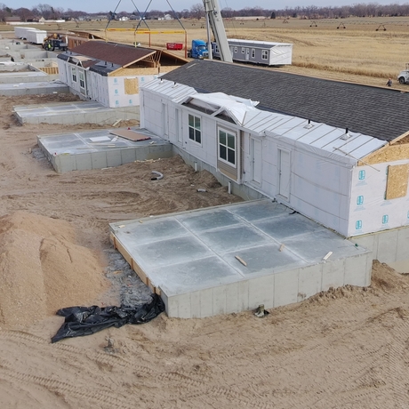 A row of single story houses under construction. The houses don't have siding yet and have shingles on the roofs. A concrete pad is in front of each house and it's all surrounded by bare brown dirt.