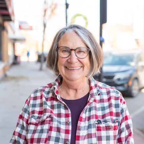 Older white woman with glasses and medium length gray hair, wearing a red and black plaid shirt, smiling, standing on a downtown sidewalk