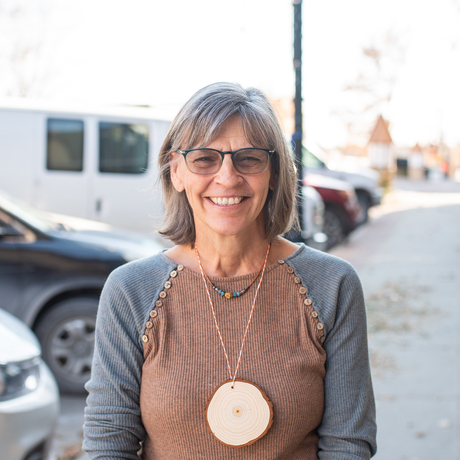 Woman with glasses and medium length gray hair wearing a brown and gray shirt, standing on a downtown sidewalk