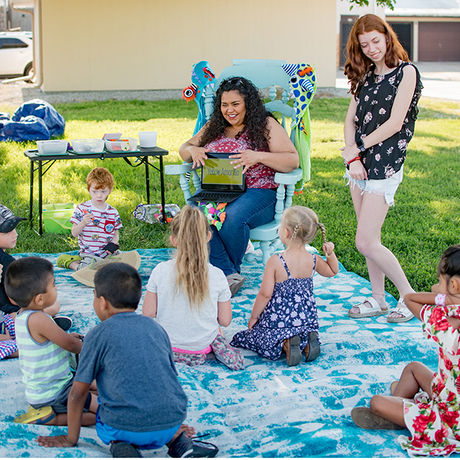 Woman sitting on a chair reading a book to children sitting in front of her on a blanket