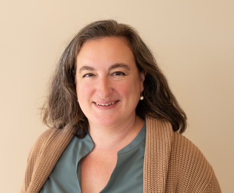 Woman with brown hair, wearing a blue-green shirt with a brown sweater over it