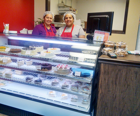Two women standing behind a light-up display case full of cakes