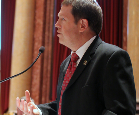 Former Iowa State Senator Tod Bowman, standing at a microphone in a black suit jacket, red tie, and white shirt. 