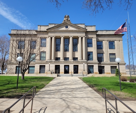 County government building with a sidewalk and green grass and flag pole in the foreground, and a blue sky. 