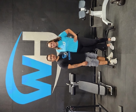 A hispanic male wearing a black shirt and grey shorts stands next to a hispanic female wearing a blue shirt and black pants inside a gym with gym equipment around them