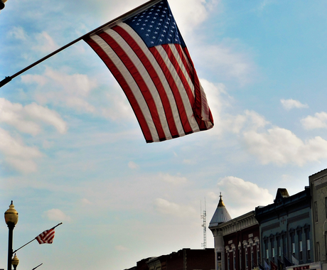 An American flag waves, suspended from a street lamp on a small town Main Street. The sky is blue with fluffy, white clouds, and the street is lined with historic brick buildings.