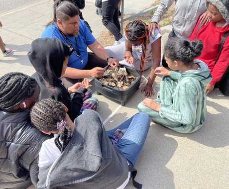 Hispanic male wearing blue shirt in center of image shows items in bin to African American kids sitting around him on concrete stairs near a sidewalk