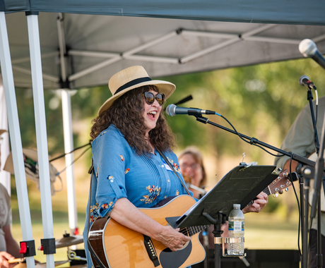 Woman in blue button up short-sleeved top with flower prints, wearing a tan sunhat with a black ribbon. Woman has sunglasses on, brown curly hair to her shoulders, and is singing into a microphone, strumming a guitar.