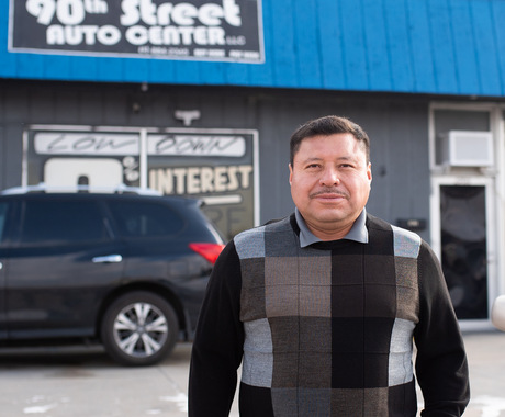 A dark-haired man wearing a black, brown, and grey checkered sweater looks at the camera in front of a building with a bright blue awning. The sign on the building says "90th Street Auto Center."