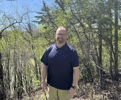 Man with thin hair on his head, dark glasses, and facial hair, wearing a blue polo shirt and khakis stands in a wooded area, smiling at the camera.