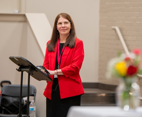 Woman with dark red hair wears a black shirt and black pants with a red suit jacket over it. She is standing at a podium with painted white brick walls behind her.