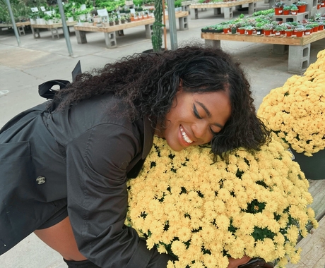 African American woman wearing a long sleeve jacket and high black boots hugs a pot of yellow flowers in a greenhouse