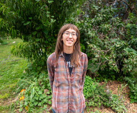 Woman standing in front of a tree/bush. Woman has brown hair with bangs, glasses, a reddish plaid shirt and is smiling directly at the camera.